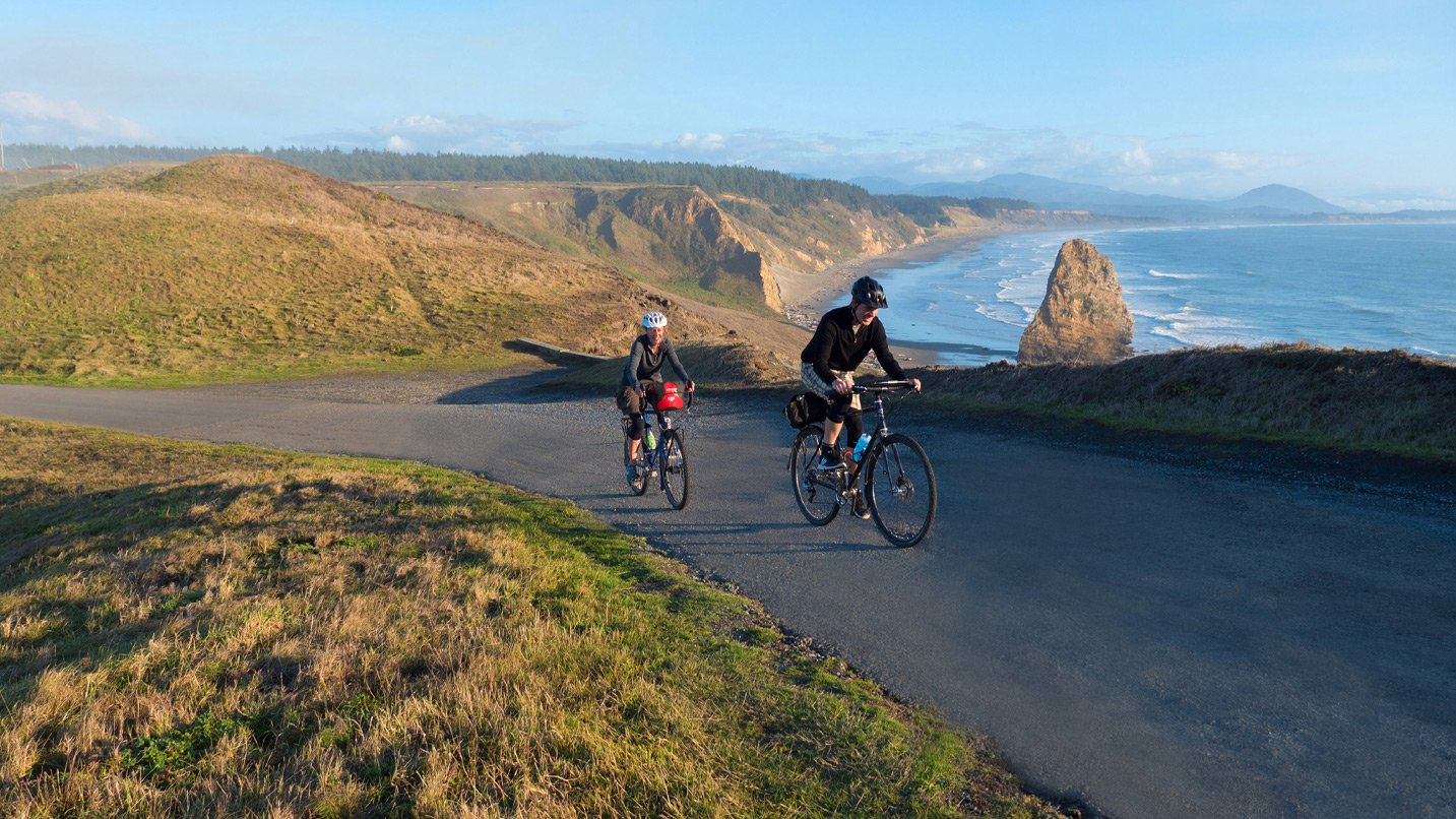 Two cyclists ride up a hill with a scenic coastal backdrop.
