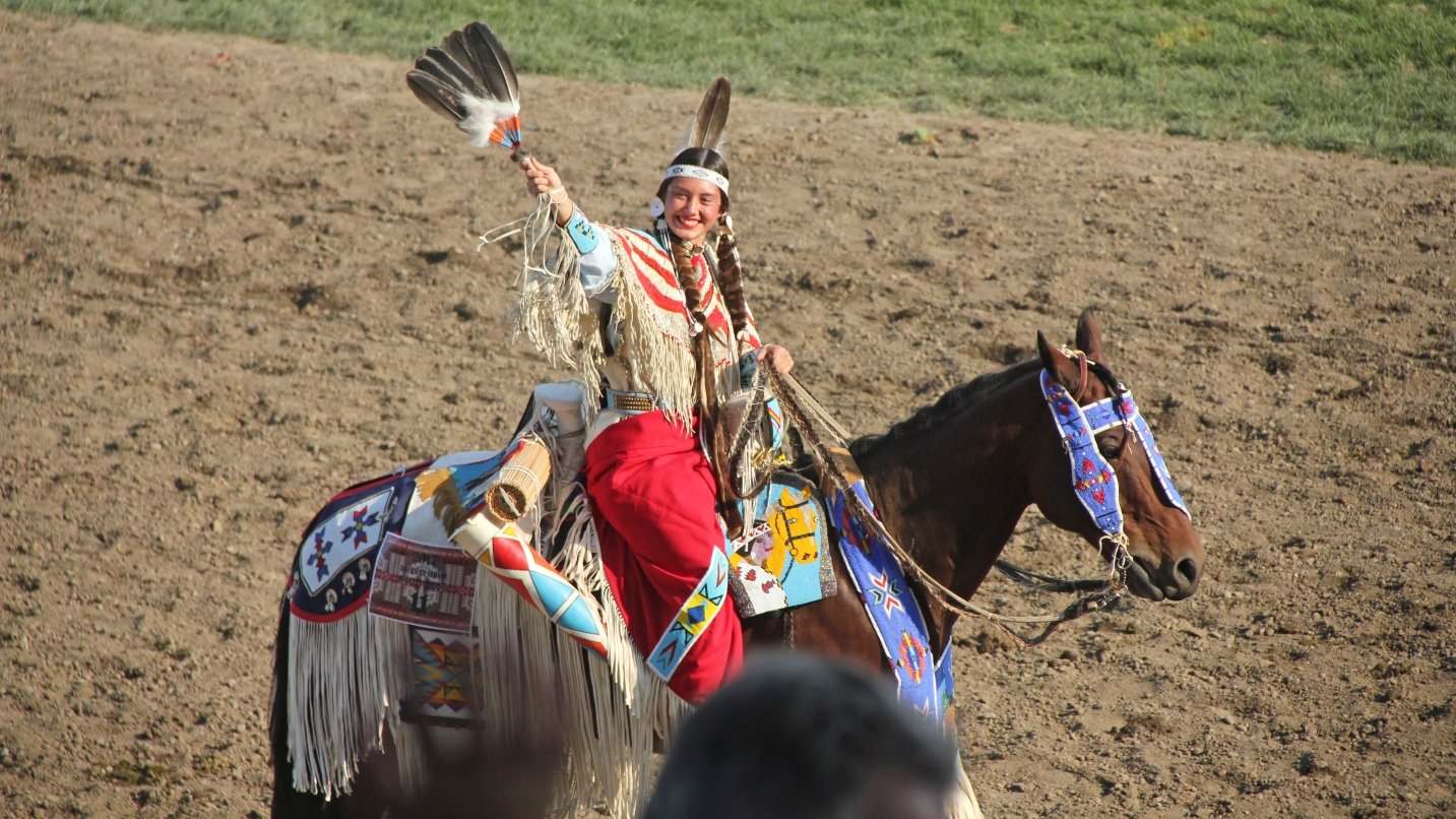 Woman in native regalia smiles and waves on a horse