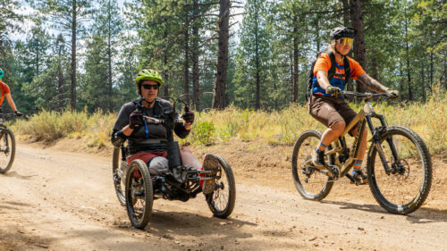 Two cyclists on two-wheeled mountain bikes and one cyclist in a 3-wheeled adaptive bike on a dirt road.
