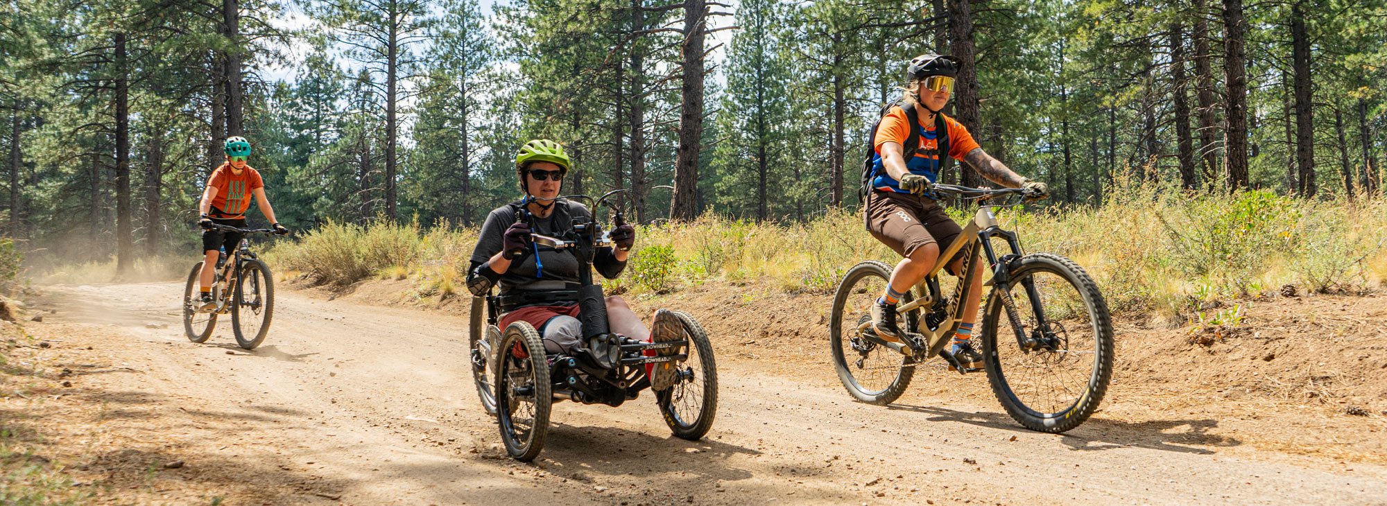 Two cyclists on two-wheeled mountain bikes and one cyclist in a 3-wheeled adaptive bike on a dirt road.
