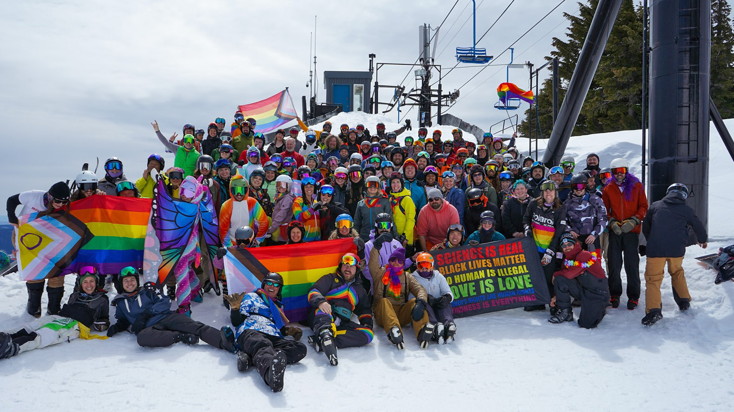 A large group of people in snow gear pose for a group photo with several rainbow and LGBT+ flags by outdoor ski lifts.