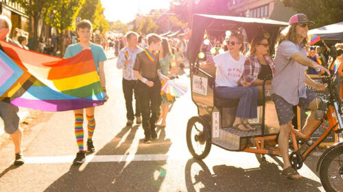 A group of festival goers at a Pride festival. Some hold up a LGBT flag, others ride a taxi bike.