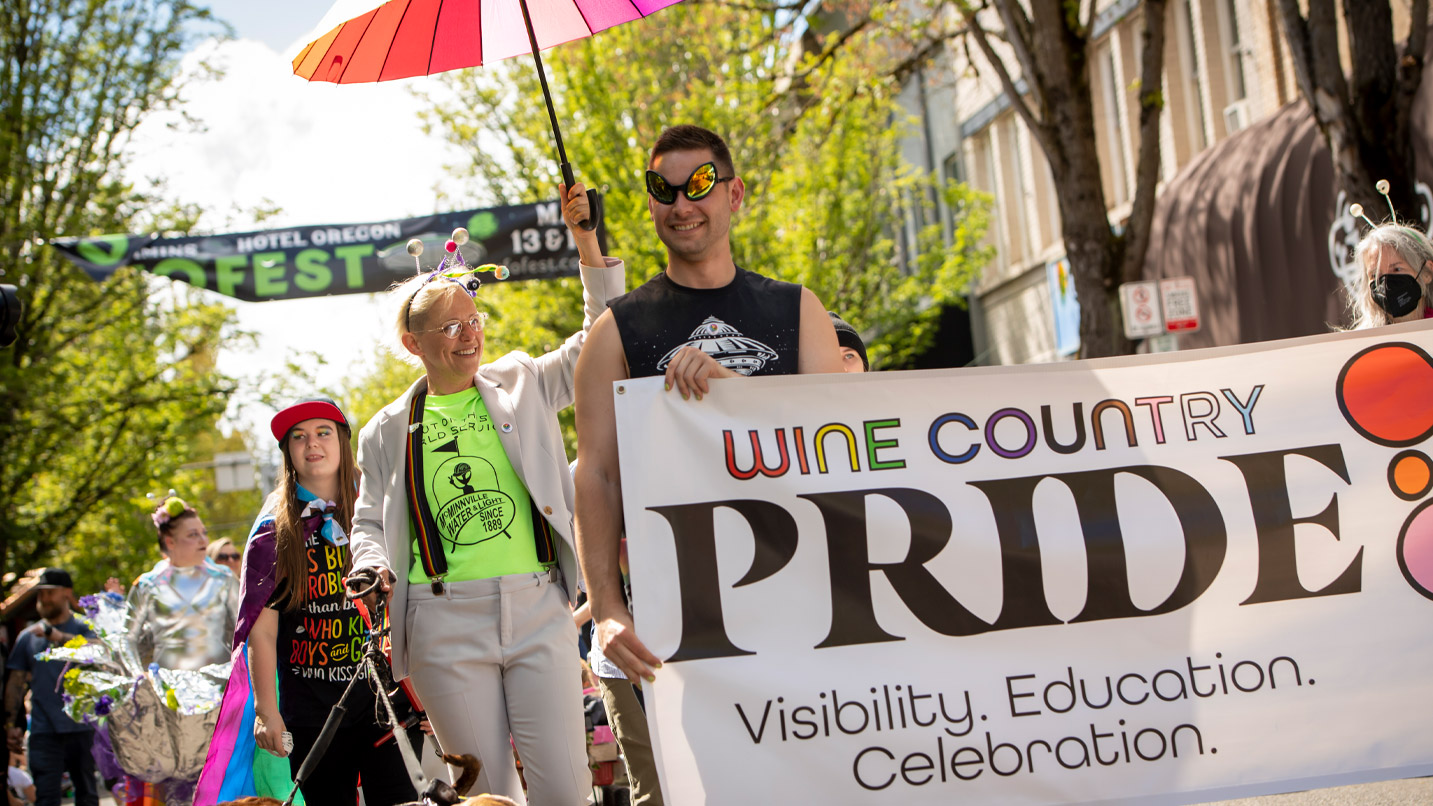 People in bright clothing and accessories march down a main street with a banner reading "Wine Country Pride"