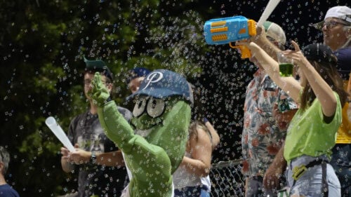 A woman shoots bubbles out of a bubble machine as a pickle mascot and fans watch.