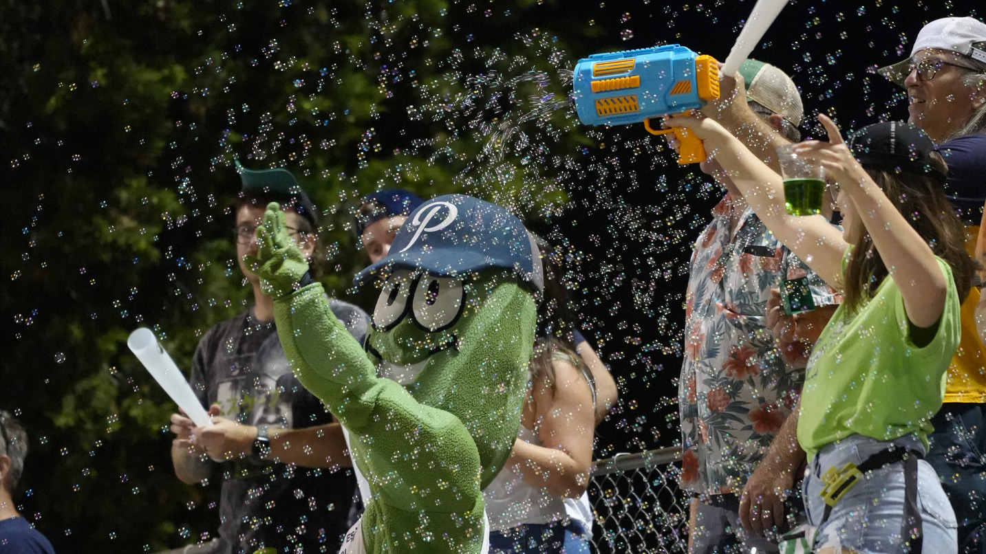 A woman shoots bubbles out of a bubble machine as a pickle mascot and fans watch.