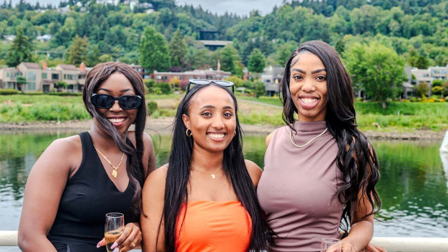 Three women stand at the top of a boat holding wine glasses.
