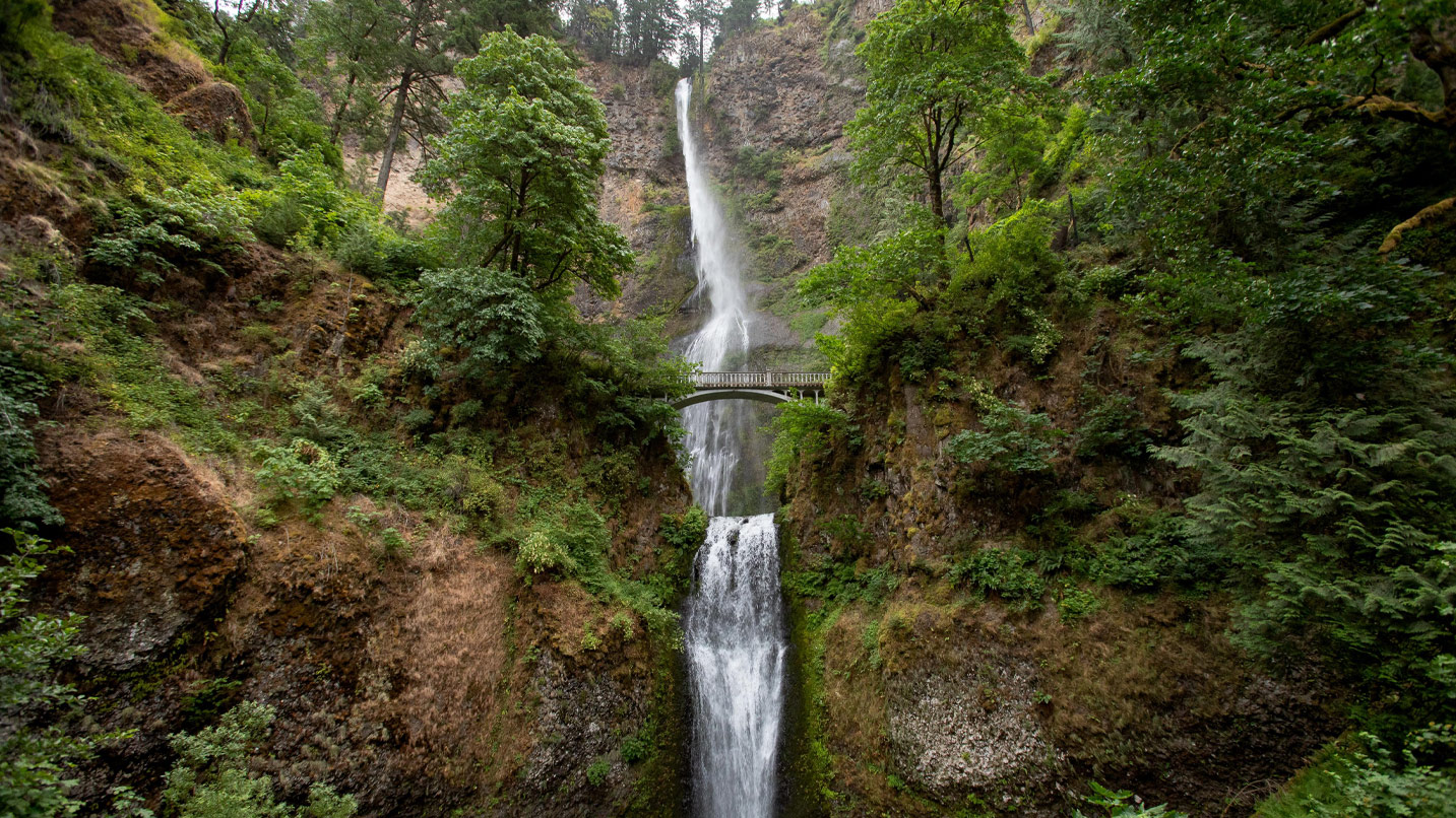 A two-tiered waterfall passes under a stone pedestrian bridge.