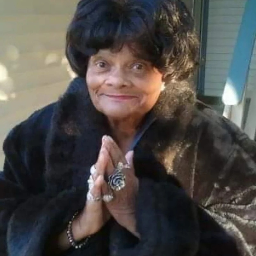 Older black woman with black hair smiles, wears a black coat and holds hands together