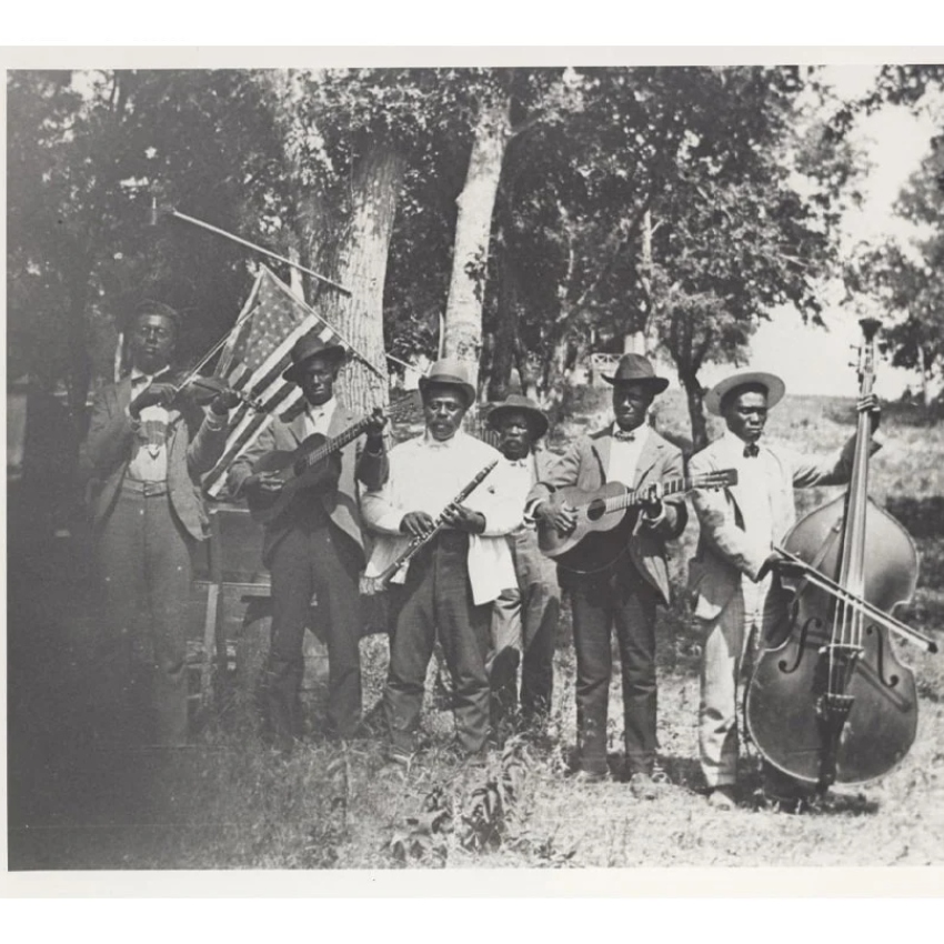 Black and white image of six Black men with hats holding musical instruments in a field