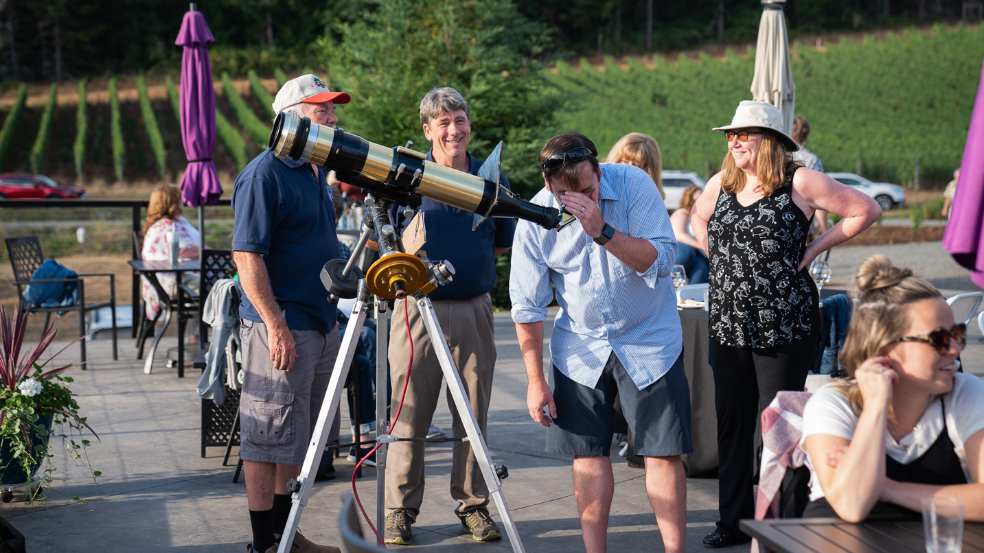 A group of people on a vineyard's patio with a telescope set up.