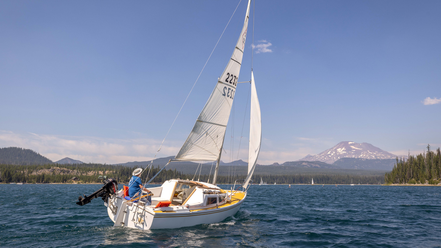 A man in a sailboat on calm waters looking towards a mountain.