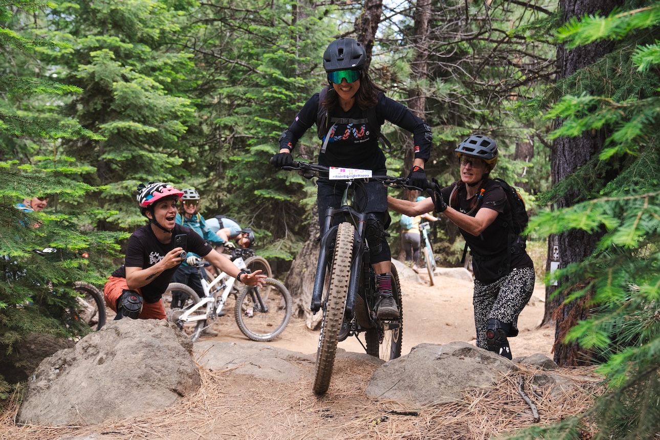 A pair of female mountain bikers support a biker as she attempts to ascend a rock feature.