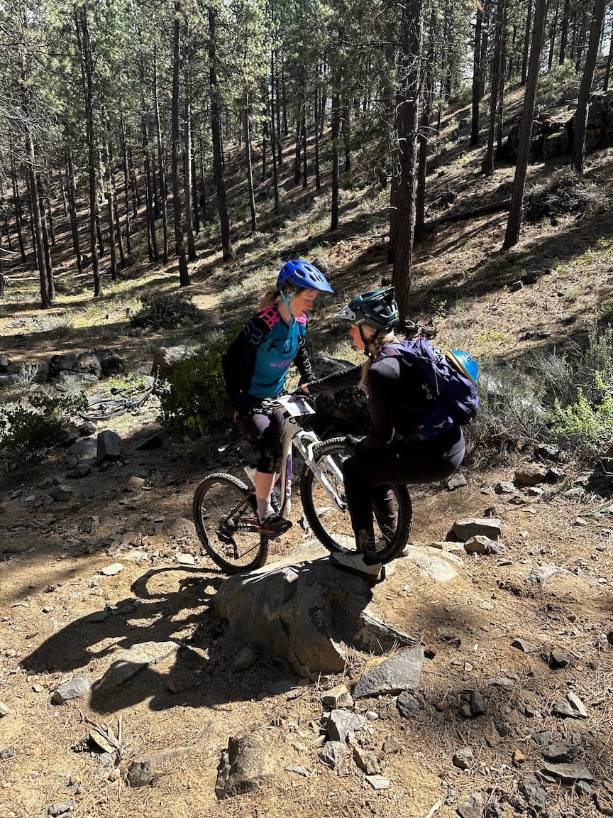 A mountain biker pauses on a rock with support from their coach.