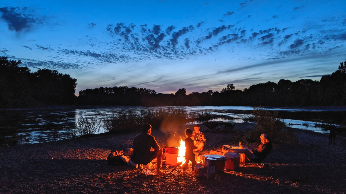 People sit around a campfire under a dark blue sky and body of water