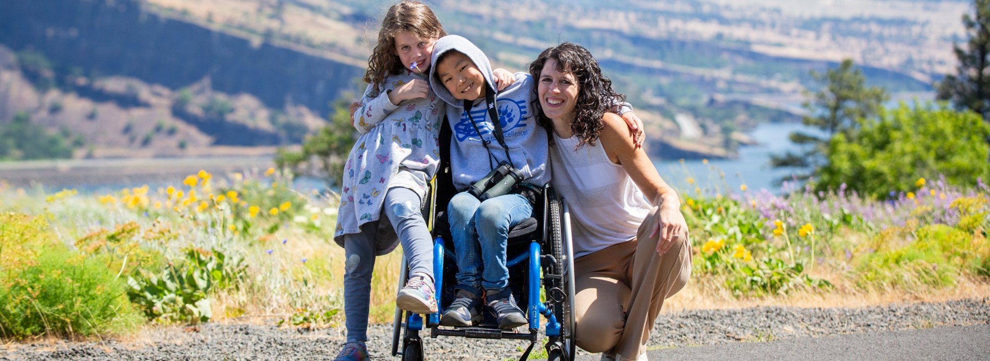 Woman, boy in wheelchair and girl pose on paved trail with wildflowers and river gorge in background