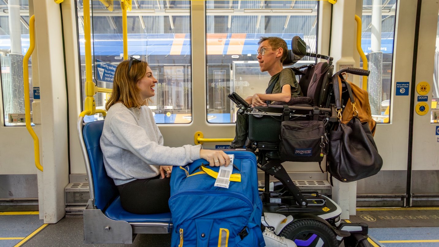 Woman sits on light-rail train holding blue suitcase, across from man in wheelchair