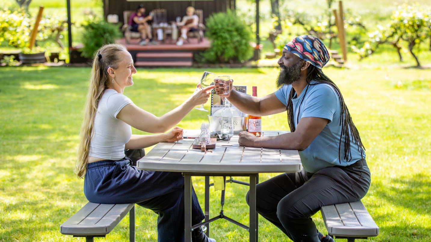 Woman and man toast wine glasses at a picnic table with grass and a stage in background