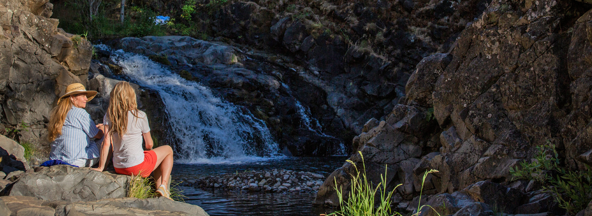 Two women sitting on a rock by a small river waterfall.