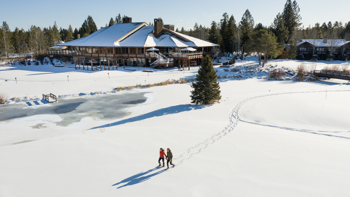 Two people walk through the snow with the lodge in the background.
