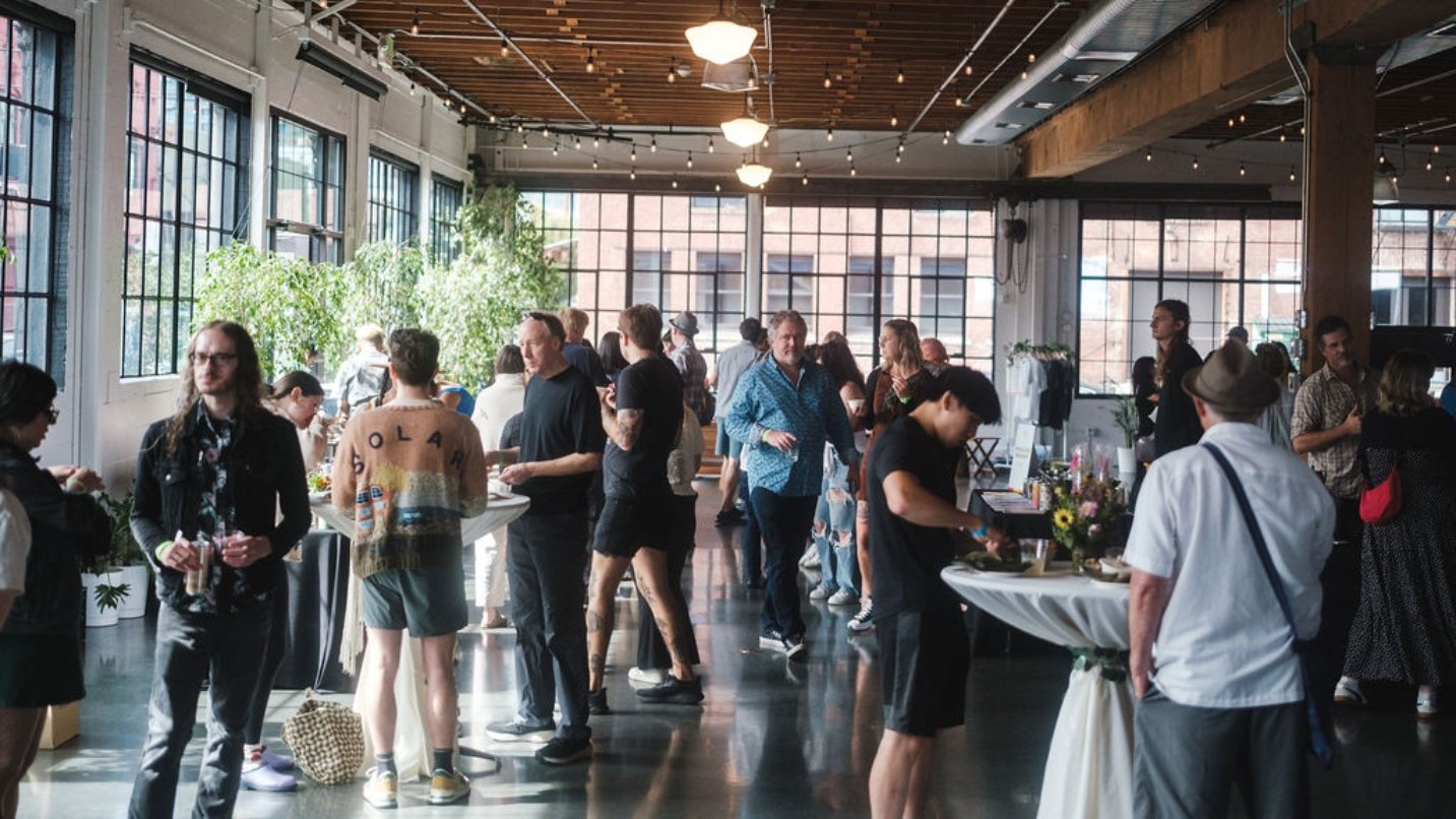 People stand around tables sampling food and socializing in a building with high ceilings, large windows and cool lighting