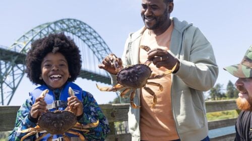 Man and little boy holding large crabs and smiling, with bridge in background