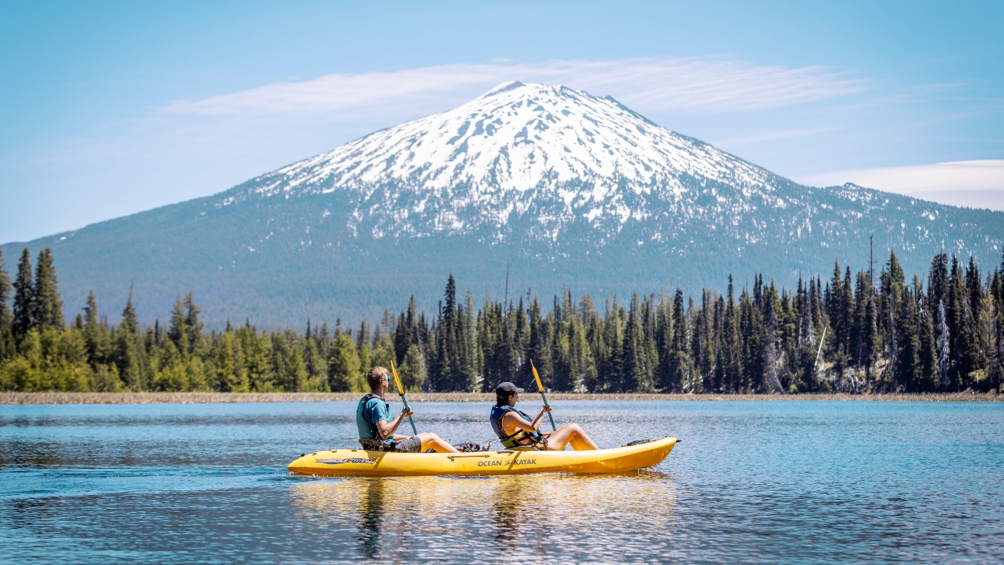 Woman and man paddle a kayak on lake with trees and snow-capped mountain in background