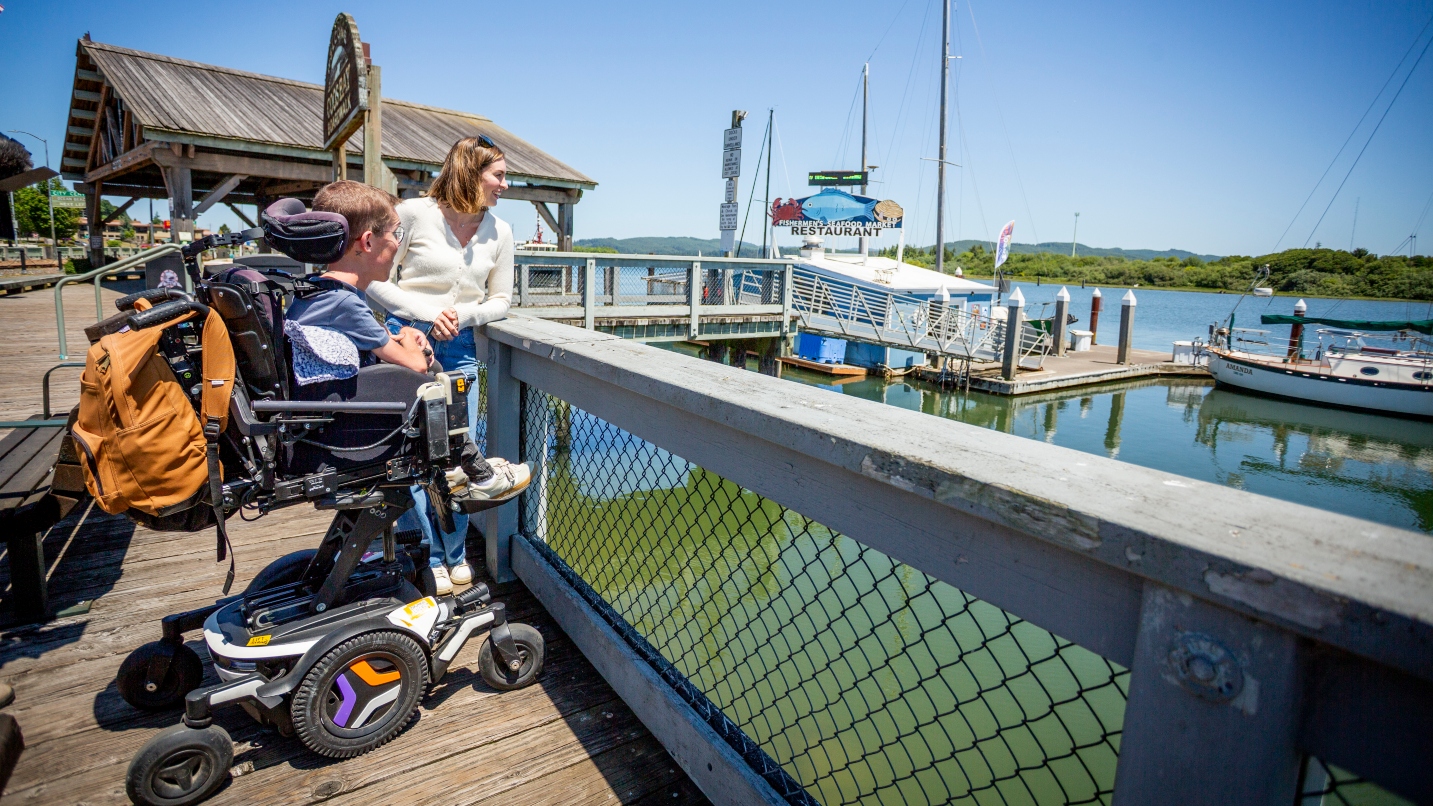Woman looks at marina with man in wheelchair on boardwalk
