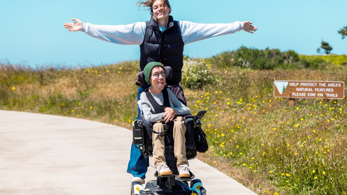 Woman smiles and stands behind man smiling in wheelchair, with grasses next to paved path
