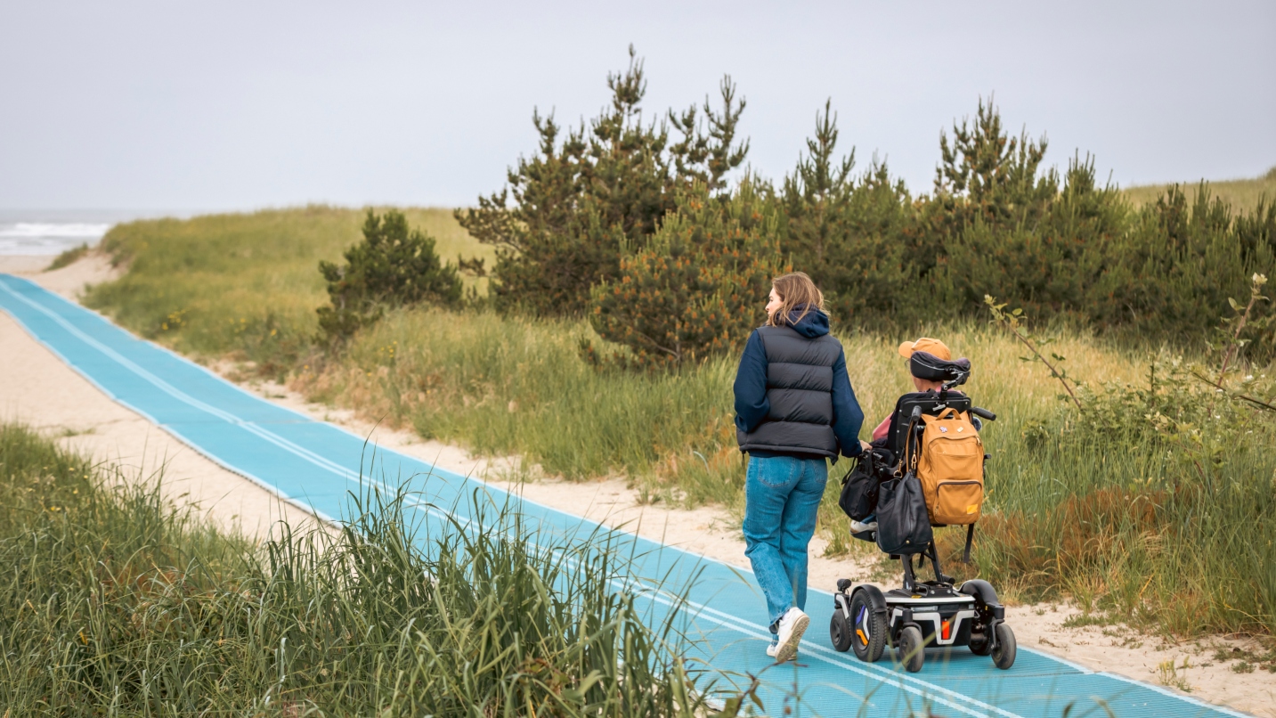 Woman walks along long blue mat with man in wheelchair, with vegetation and sand on either side