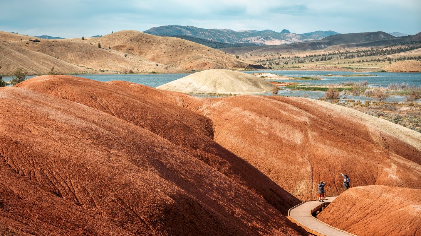 Two people on paved boardwalk in foreground with reddish and brown hills in background