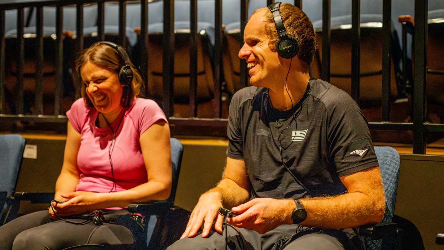 Man and woman sit in theater seats with headphones on and smile
