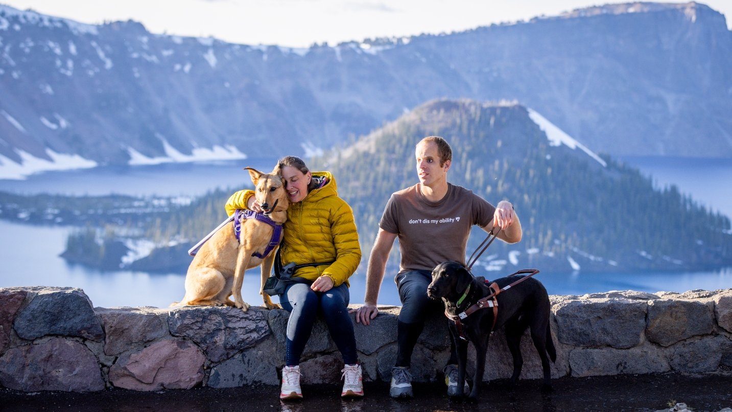 Man and woman sit on rock wall, holding dogs, with large lake and island in background