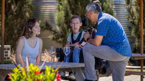 Man puts leg on picnic table and shows bottle of wine to man and woman, with wine glasses and flowers in foreground