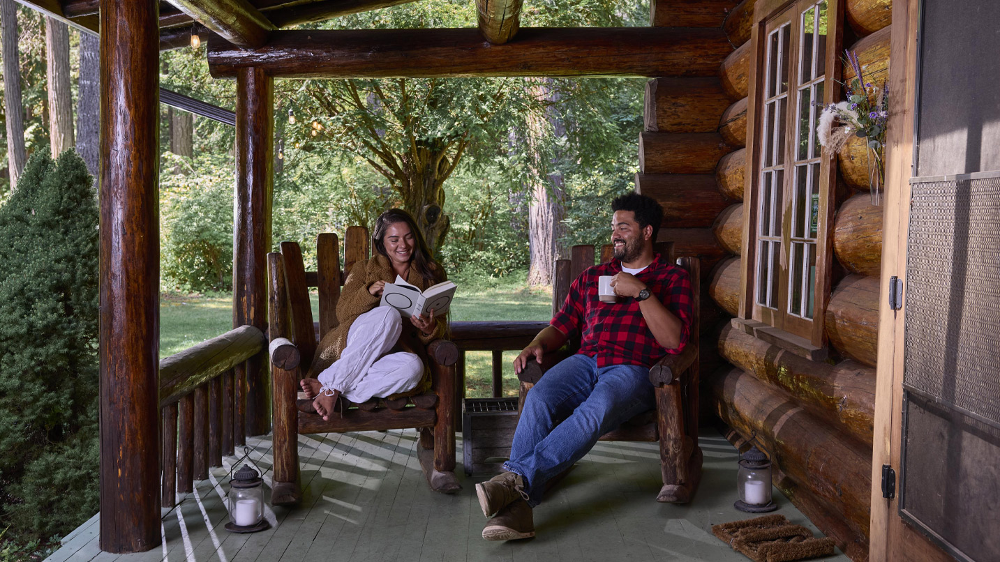 Two people drinking coffee on a wooden cabin's covered front porch.