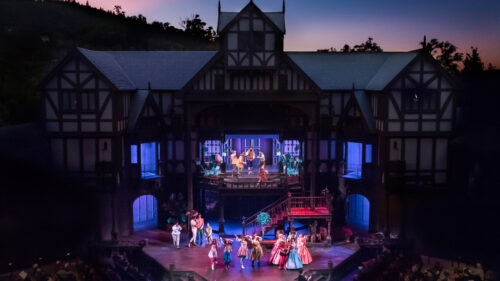 A play performed in an outdoor stage, with the backdrop set of a old English style building.