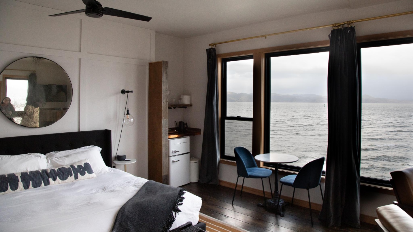 Hotel room with bed, chairs, table, fridge, curtains and large window opening up to riverfront view