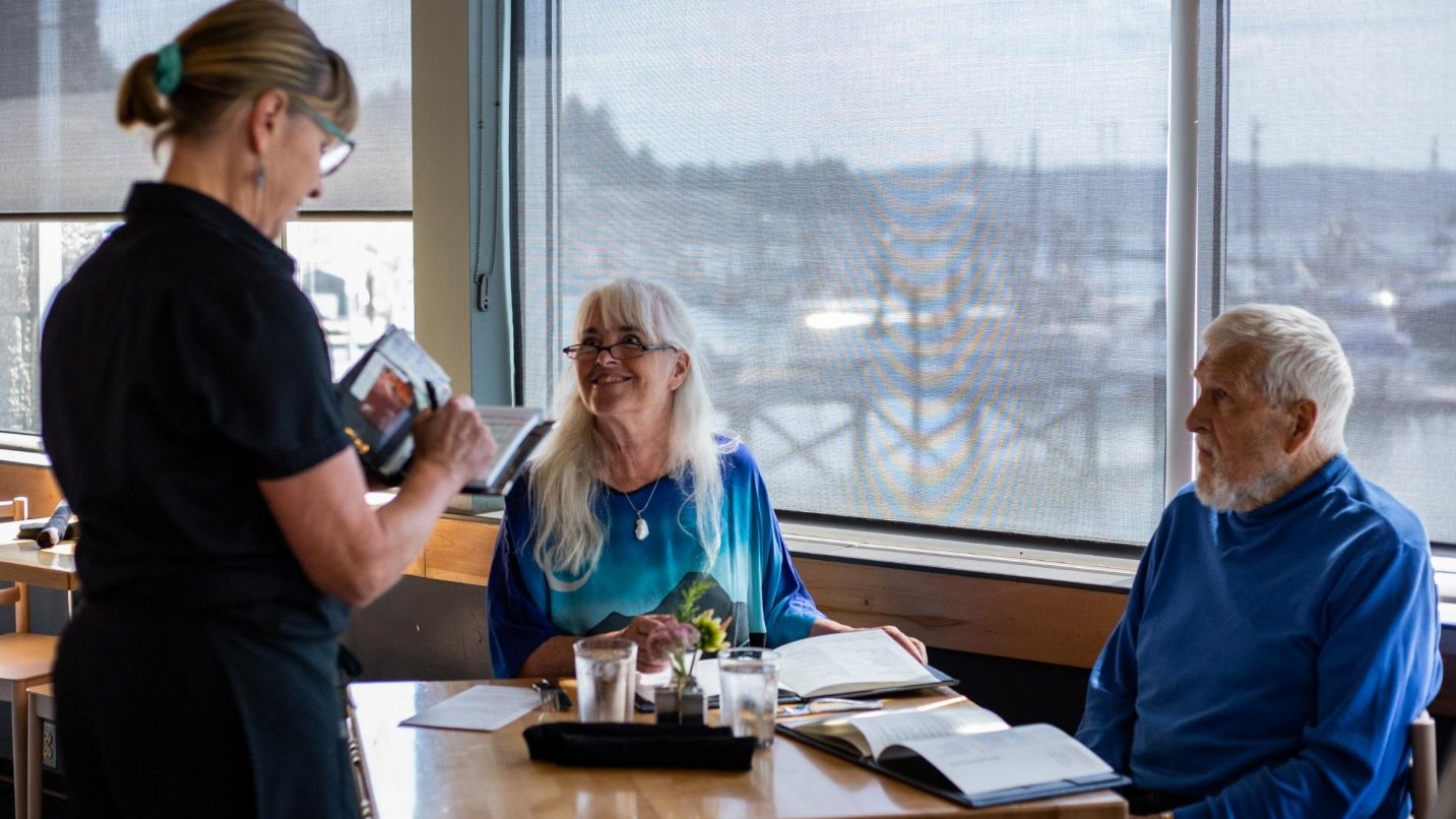 Man and woman with grey hair sit at restaurant table with large windows overlooking a bay, waitress standing in front of them