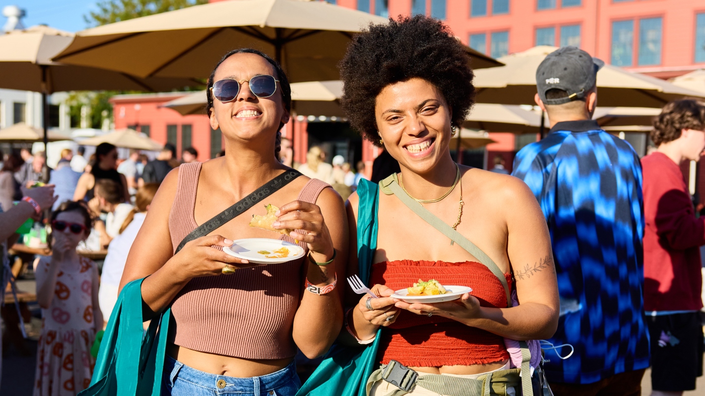 Two women smile at camera while holding plates of food in sunshine