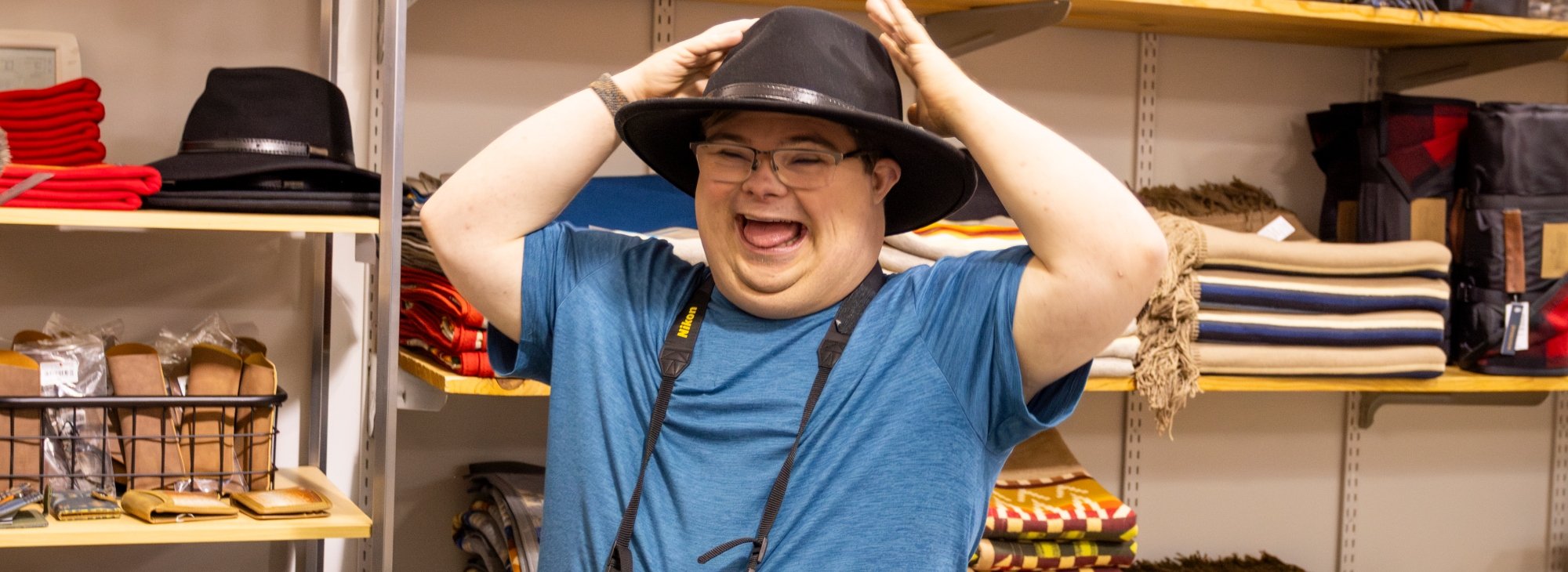 Man wearing blue shirt smiles broadly and holds hands up to his wide-brimmed black hat