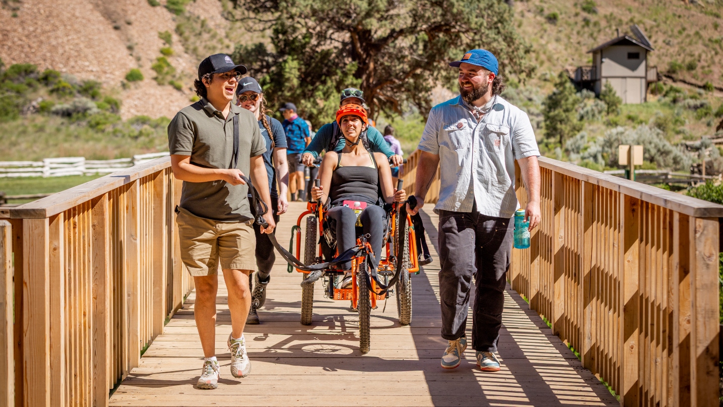 Several people walk across a wooden boardwalk holding harnesses to an all-terrain wheelchair with woman sitting in it