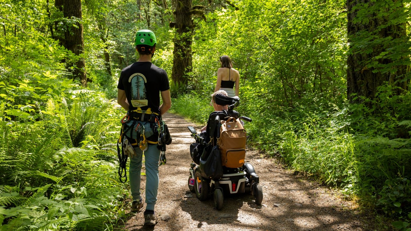 Two people walk and another person uses a wheelchair along a dirt trail in a forest