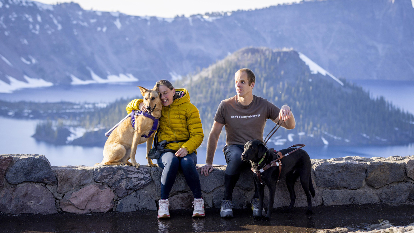 Two people and their service dogs sit against a backdrop of Crater Lake.