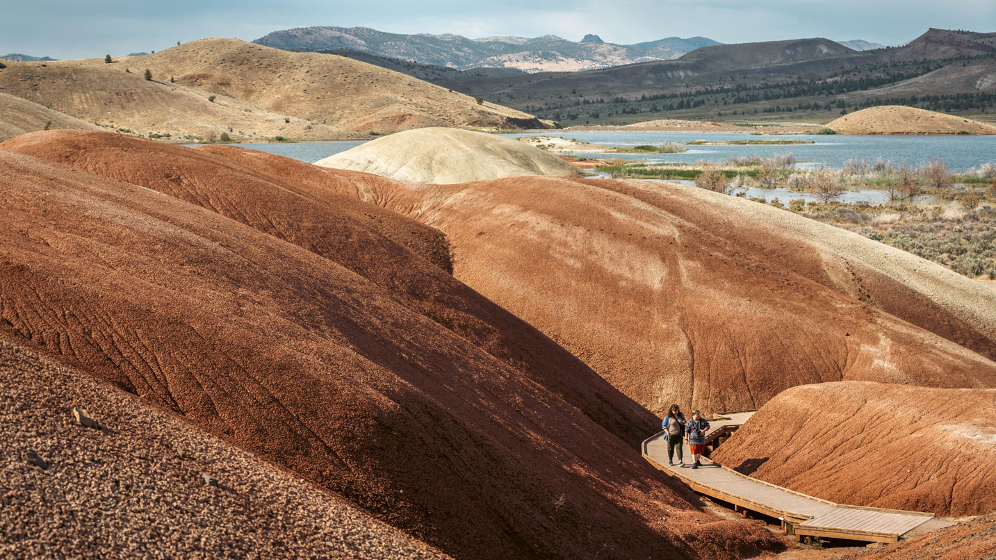 Two people walk on a wood boardwalk through red claystone hills.