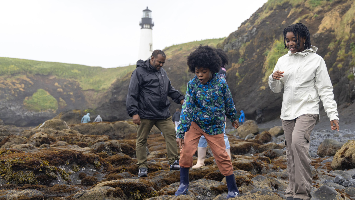 A family walks along a rocky shore near a lighthouse.