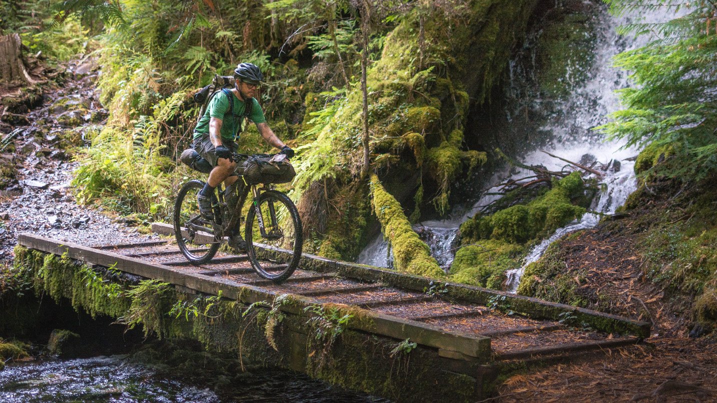 A mountain biker on a short rustic bridge passing a waterfall.