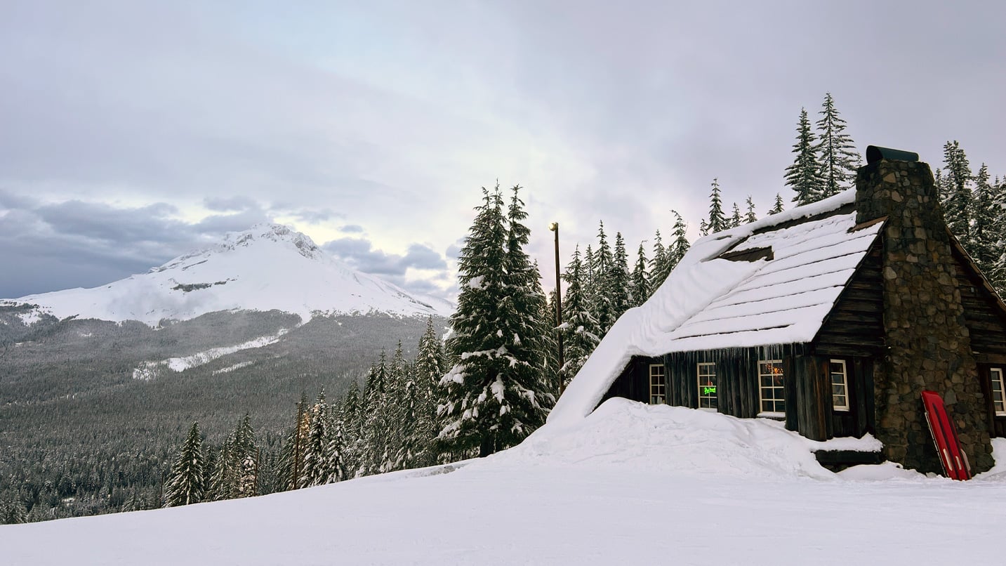 Snow covers a historic wooden cabin with a view of a mountain in the distance.