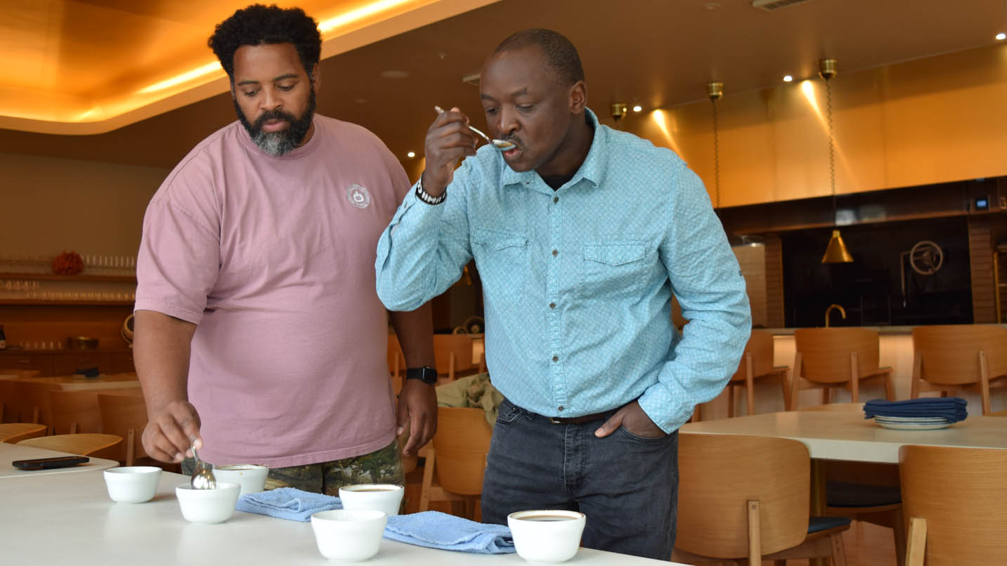 Two men hold spoons that they are dipping into white mugs on table and sipping from, with restaurant tables and chairs in background