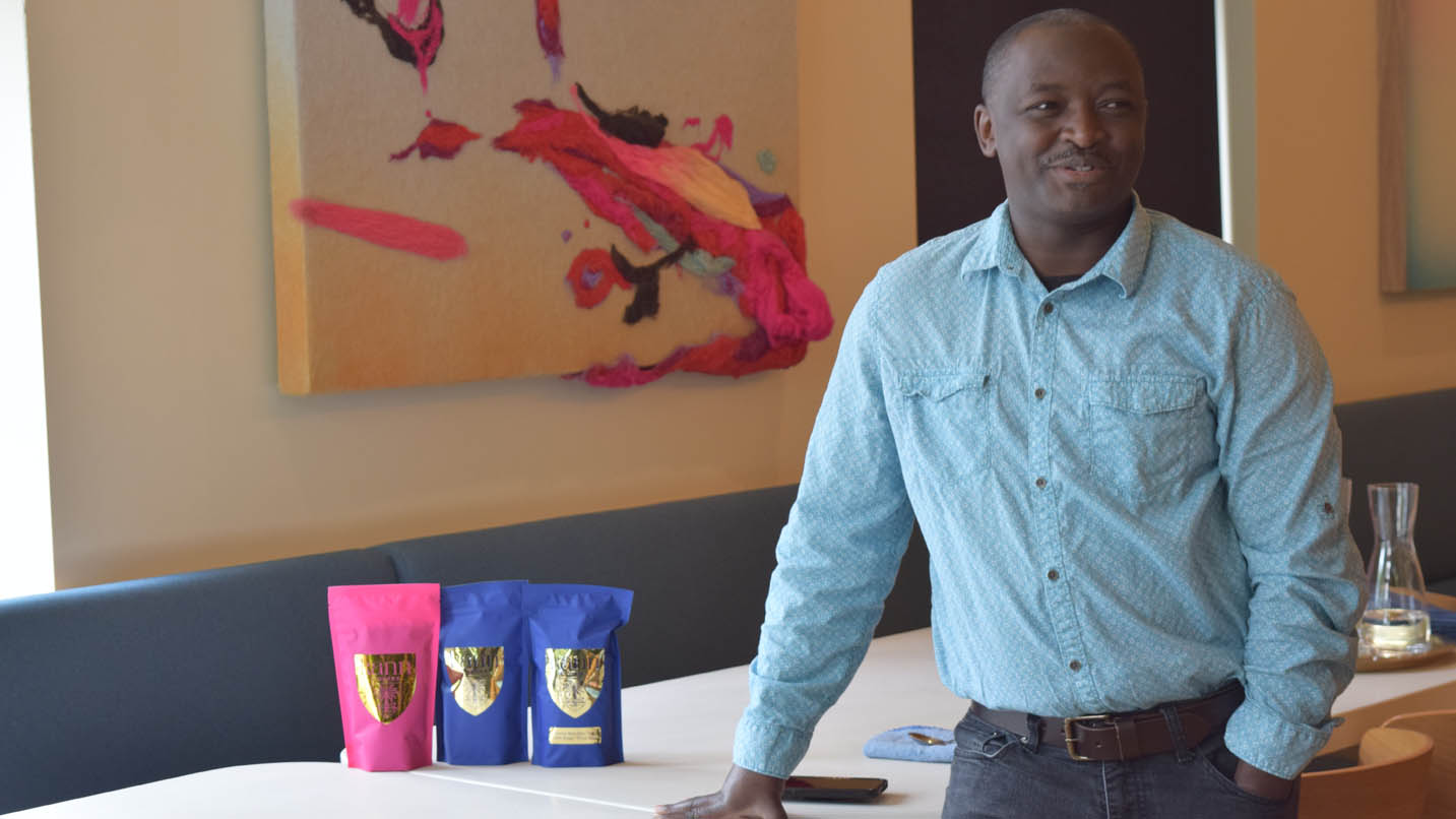 Man smiles with hand in pocket, other hand on table and colorful bags of coffee on table. Modern art painting in background.