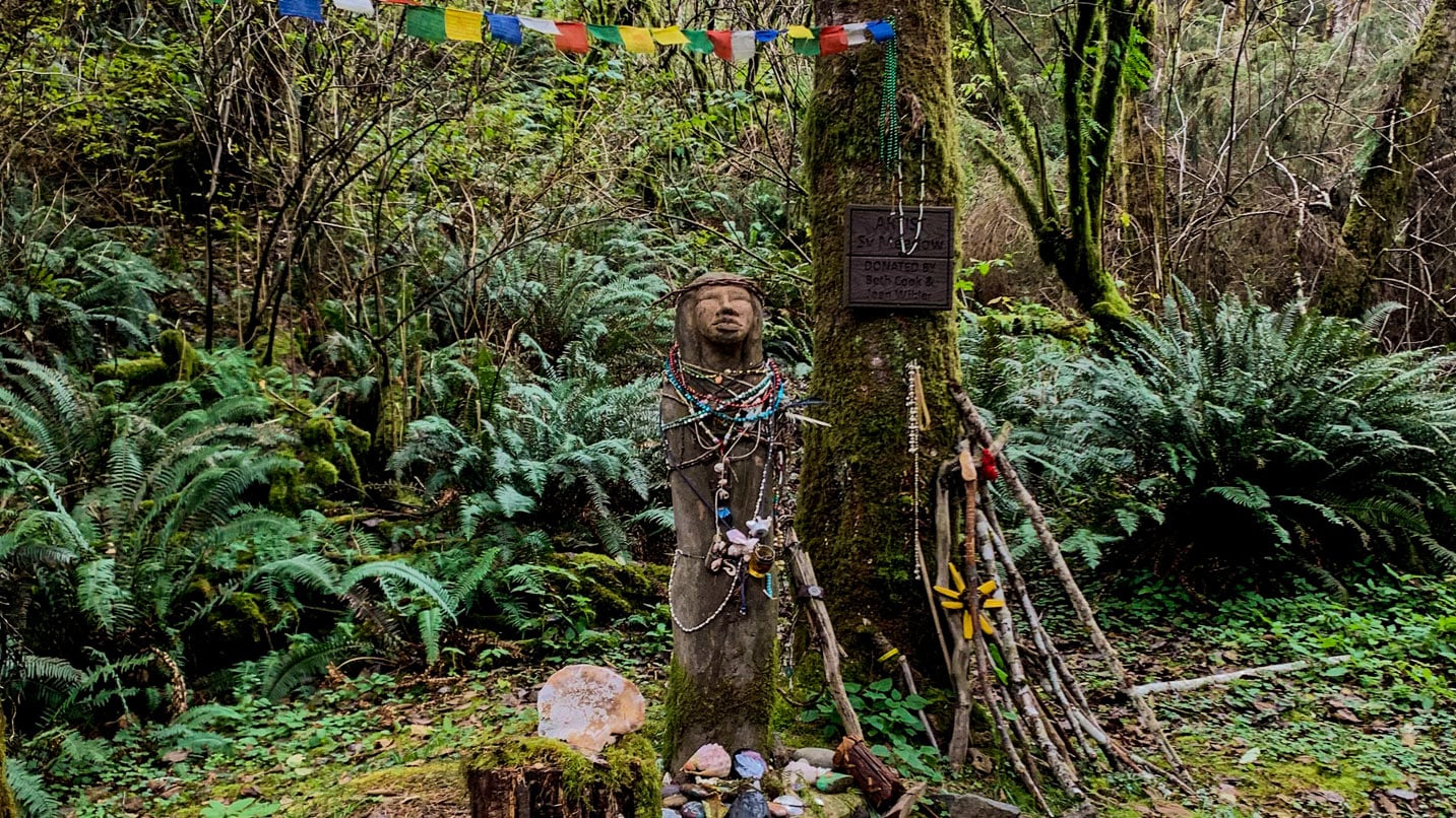A small, wodden statue of an Indigenous figure decorated with small stones, beads and other artifacts.