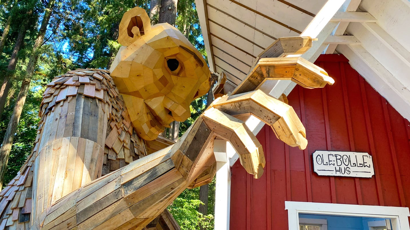 A large outdoor wooden sculpture of a troll looks down into a set-designed house.